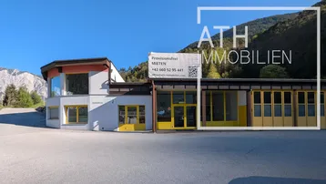 Außenansicht eines Gewerbegebäudes mit großem Parkplatz, umgeben von einer malerischen Berglandschaft und klarem Himmel.