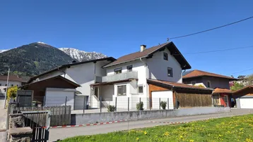 Weißes Wohnhaus mit braunem Dach und Balkon, umgeben von grünen Wiesen und Bergen.