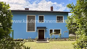 Blaues Einfamilienhaus mit gepflegtem Vorgarten und grüner Umgebung unter blauem Himmel.