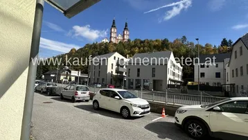 Blick auf eine Straße mit geparkten Autos, modernen Gebäuden und einer Kirche auf einem bewaldeten Hügel im Hintergrund.
