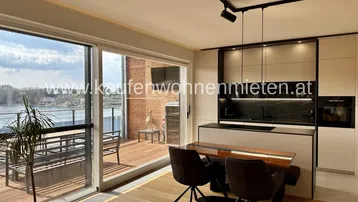 Moderner offener Wohn-Essbereich mit integrierter Küche, Holzboden und Zugang zu einer großen Terrasse mit Seeblick.