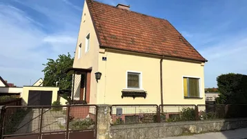 Gelbes Einfamilienhaus mit rotem Ziegeldach und Vorgarten, umgeben von einem Zaun.