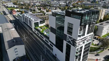 Luftaufnahme eines modernen Gebäudekomplexes mit Glasfassaden und Gründächern neben Bahngleisen in städtischer Umgebung.
