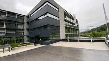 Modernes Gebäude mit dunkler Fassade, Balkonen und Ladestationen für Elektrofahrzeuge im Vordergrund.