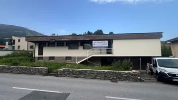 Gebäudeansicht mit einem 'Zu verkaufen'-Schild, umgeben von Grünflächen und mit Bergblick im Hintergrund.