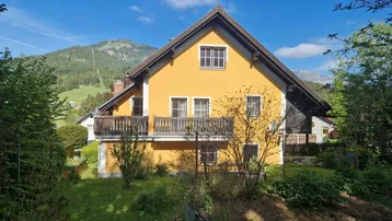 Gelbes Haus mit Satteldach und Balkon, umgeben von grüner Natur und Bergen im Hintergrund.