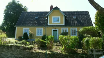 Gelbes Landhaus mit grünem Eingang und gepflegter Hecke im Sonnenschein.