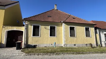 Gelbe Fassade eines traditionellen Hauses mit Ziegeldach und gepflastertem Vorplatz.