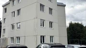 Mehrstöckiges Wohngebäude mit mehreren Fenstern und geparkten Autos davor unter bewölktem Himmel.