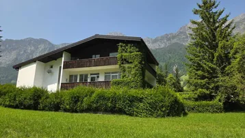 Das Haus präsentiert sich mit einem Balkon und ist umgeben von einer gepflegten Hecke und grünen Wiesen mit Bergblick.