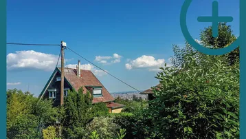 Haus mit Satteldach und weitem Blick über die Stadt, umgeben von üppigem Grün.