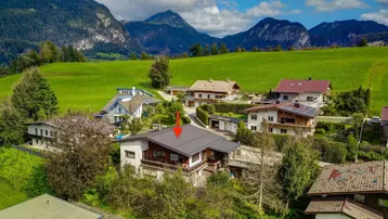 Luftaufnahme eines Hauses in ländlicher Umgebung mit grünen Wiesen und majestätischen Bergen im Hintergrund.