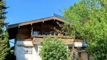 Traditionelles Holzhaus mit Balkon und Solaranlage auf dem Dach, umgeben von Bäumen.