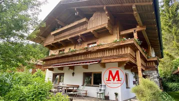Traditionelles Holzhaus im alpinen Stil mit Terrasse und Gartenbereich, umgeben von üppigem Grün.