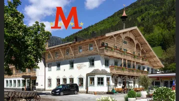 Traditionelles Gasthaus mit Holzbalkonen und Blumenkästen, umgeben von grünen Bergen unter blauem Himmel.