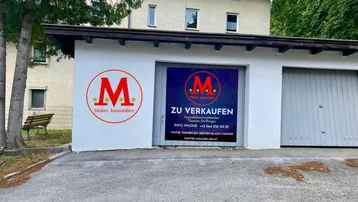 Weiße Garage mit zwei Toren und einem 'Zu verkaufen'-Schild, davor eine asphaltierte Fläche und im Hintergrund ein Wohngebäude.