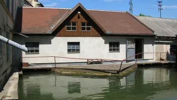 Historisches Gebäude am Wasser mit braunem Ziegeldach und Holzgiebel, umgeben von einer Holzplattform.