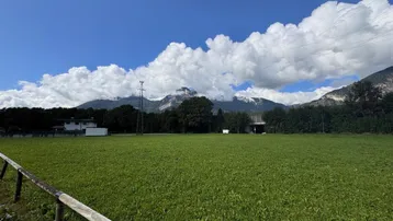 Weitläufige grüne Wiese mit majestätischen Bergen im Hintergrund unter einem strahlend blauen Himmel.