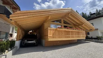 Modernes Holzchalet mit Carport und Balkon in alpiner Umgebung unter blauem Himmel.