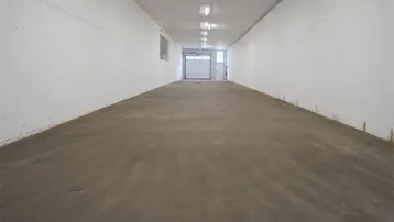 Langer, leerer Lagerraum mit Betonboden und Leuchtstoffröhrenbeleuchtung, der zu einem Garagentor führt.