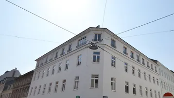 Weißes Eckgebäude mit vielen Fenstern an einer sonnigen Straße, umgeben von parkenden Autos.