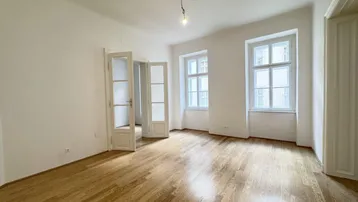 Helles Zimmer mit Holzboden und hohen Fenstern, das viel Tageslicht hereinlässt.