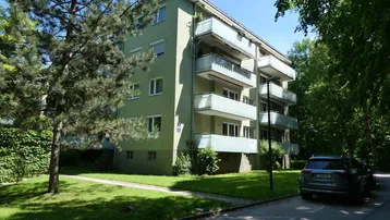 Grünes Mehrfamilienhaus mit mehreren Balkonen, umgeben von Bäumen und Grünflächen.
