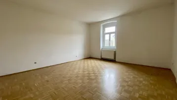 Heller, leerer Raum mit Fischgrätparkettboden und einem Fenster, das Tageslicht hereinlässt.