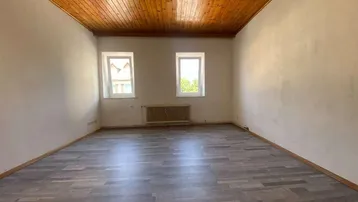 Helles Zimmer mit Holzbalkendecke, Laminatboden und zwei Fenstern, bereit zur individuellen Gestaltung.