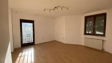 Heller Wohnraum mit Holzboden, großem Fenster und Zugang zu einem kleinen Balkon.