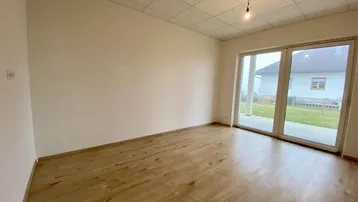Helles, leeres Zimmer mit Holzboden und großen Fenstern zum Gartenbereich.