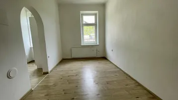 Helles, leeres Zimmer mit Holzfußboden und einem Fenster, das Tageslicht hereinlässt.