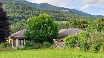 Gebäude mit großem Baum und grüner Wiese, umgeben von Bergen unter bewölktem Himmel.