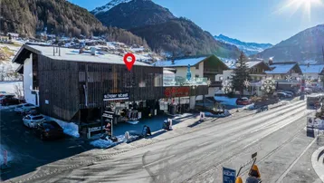 Luftaufnahme eines Gebäudes mit Holzfassade an einer Straße, umgeben von schneebedeckten Bergen unter strahlendem Sonnenschein.