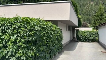 Moderne Architektur mit gepflegter Zufahrt und grüner Bepflanzung vor Bergkulisse.