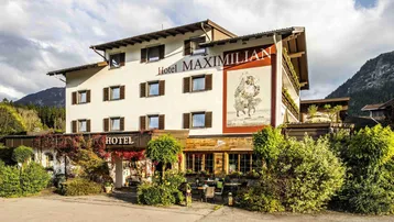 Traditionelles Hotelgebäude mit alpiner Architektur, umgeben von grüner Landschaft und Bergen im Hintergrund.