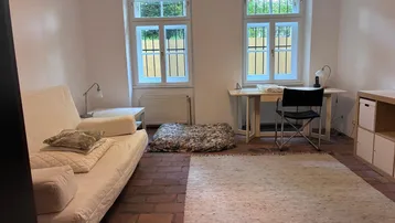 Helles Zimmer mit Ziegelboden, Schlafsofa, Schreibtisch und zwei Fenstern mit Blick ins Grüne.
