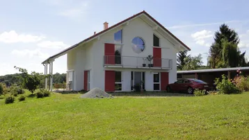 Freistehendes Einfamilienhaus mit weißer Fassade, rotem Ziegeldach und Balkon auf einem großen Grundstück.