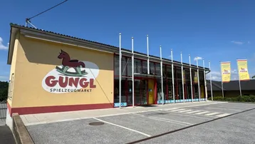 Außenansicht des Spielzeugmarktes Gungl mit großem Parkplatz und blauer Himmel.