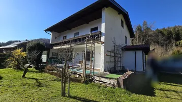 Weißes Haus mit Balkon und Terrasse, umgeben von Grünflächen und Bäumen unter blauem Himmel.