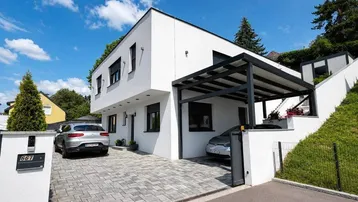 Modernes Einfamilienhaus mit weißer Fassade, Carport und gepflasterter Auffahrt unter blauem Himmel.