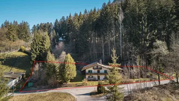 Luftaufnahme eines traditionellen Hauses mit roter Grundstücksumrandung, umgeben von Wald und Bergen.