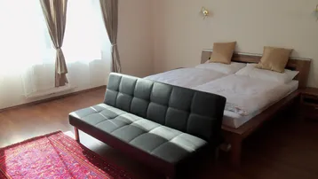 Helles Schlafzimmer mit Holzboden, Doppelbett und einem kleinen Sofa vor dem Fenster.