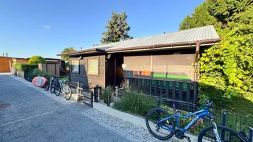 Kleines Holzhaus mit Vorgarten und Fahrrädern, umgeben von Grün und einem Kiesweg.
