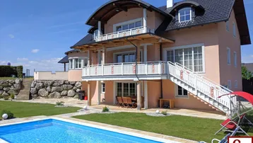 Großzügiges Einfamilienhaus mit weitläufigem Garten, privatem Pool und mehreren Balkonen unter blauem Himmel.
