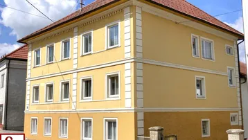 Historisches Gebäude mit gelber Fassade, weißen Fensterrahmen und rotem Ziegeldach unter blauem Himmel.
