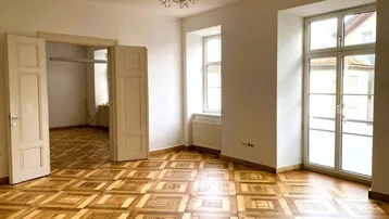 Helles Zimmer mit auffälligem Parkettboden, hohen Türen und Zugang zu einem französischen Balkon.