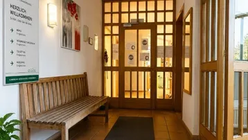 Einladender Eingangsbereich mit rustikaler Holzbank, Terrakottafliesen und großen Fenstertüren, die viel Licht hereinlassen.