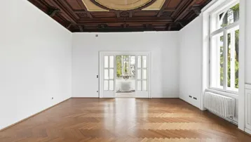 Geräumiges Zimmer mit beeindruckender Holzkassettendecke, Fischgrätparkett und großen Fenstern.