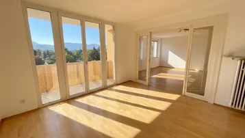 Geräumiges Wohnzimmer mit Holzboden, Zugang zum Balkon und weitem Blick auf die Berge.